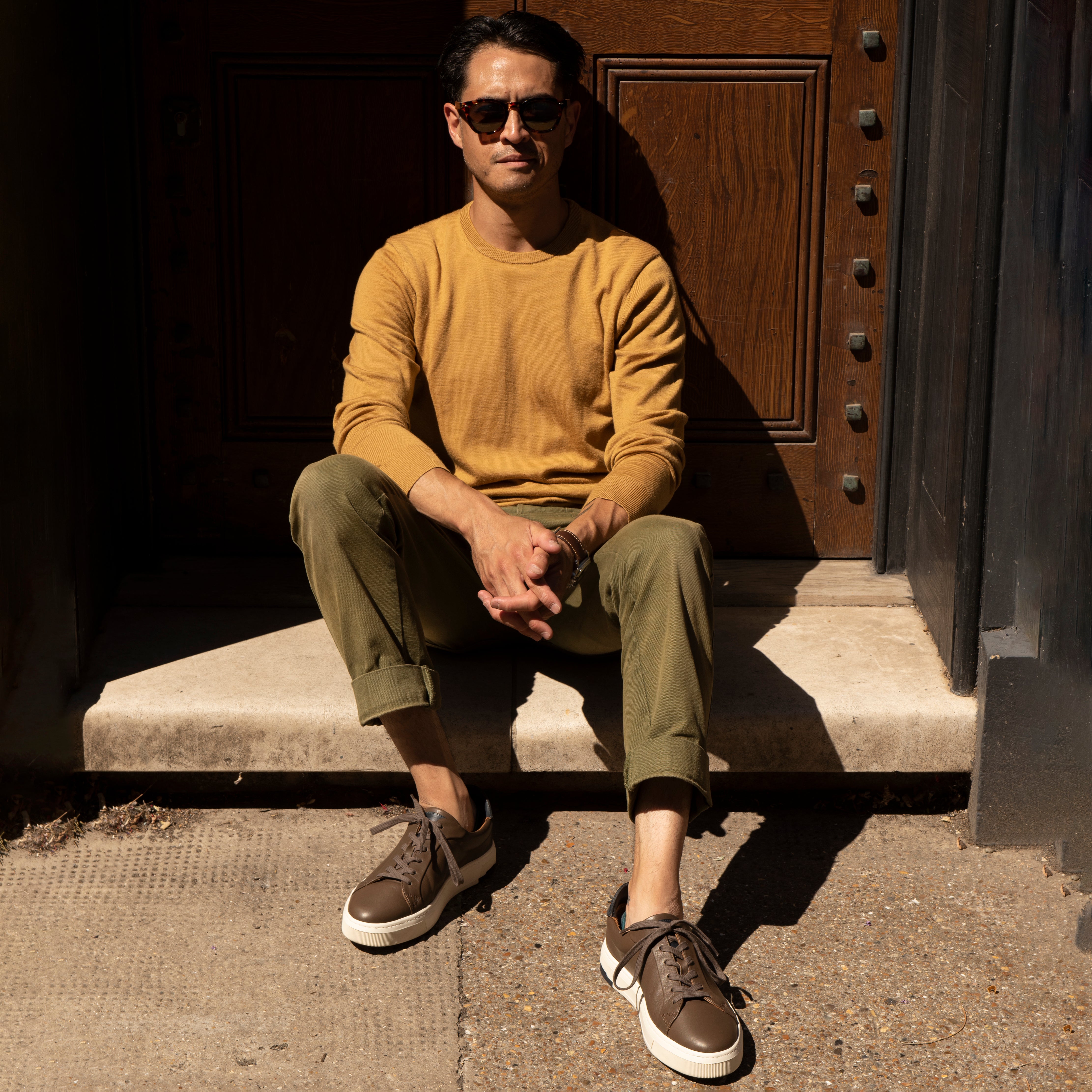Man sitting on a step wearing a mustard yellow sweater and green pants.